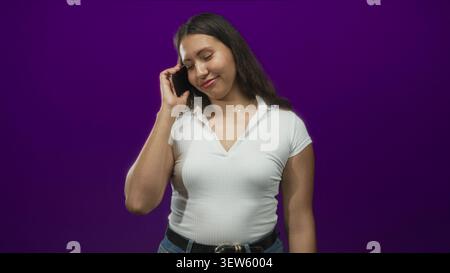 Donna che tiene il telefono all'orecchio in studio viola, sorride con la mano sollevata e indossa camicia e jeans bianchi con colletto; appagamento. Foto Stock