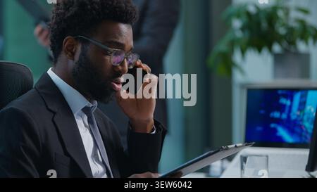 Uomo d'affari afroamericano che parla un telefono cellulare che guarda in un computer che tiene una cartella tablet con documenti che lavorano vicino al collega collega Foto Stock