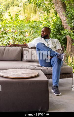 Uomo afroamericano seduto sul divano in rattan sul patio, indossa camicia, vassoio sul tavolo, spazio per fotocopie Foto Stock