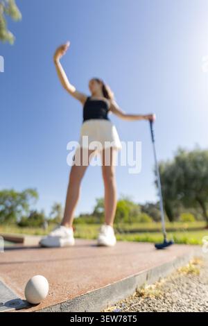 Una donna gioca a minigolf in una giornata di sole in un parco mentre fa un selfie con il telefono vicino al verde Foto Stock