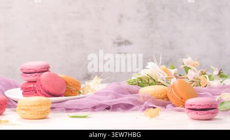 Un delicato assortimento di macaron rosa, giallo e arancione vivaci visualizzati su una superficie chiara con morbido tessuto di garza viola. Caprifoglio bianco fresco Foto Stock