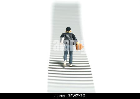 Fotografia di piccoli personaggi in miniatura. Vista posteriore di un allievo ragazzo che corre su un incrocio zebra. Isolato su sfondo bianco. Immagine Pol Foto Stock