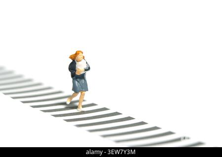 Fotografia di piccoli personaggi in miniatura. Vista frontale di una studentessa studentessa che corre su un incrocio zebra. Isolato su sfondo bianco. Immagine p Foto Stock