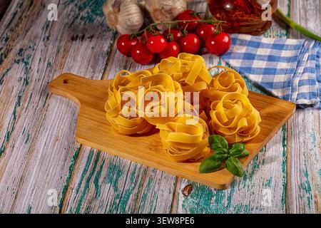 Nidi di di pasta cruda disposti su tagliere di legno con basilico fresco pomodori ciliegini e olio d'oliva. Cucina rustica con legno stagionato su Foto Stock