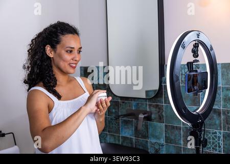 Creatrice femminile con parte superiore bianca che tiene il vaso di panna mostrando al telefono il ring light alla vanità Foto Stock