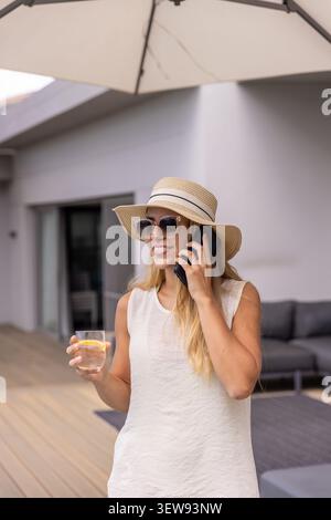 Donna che indossa un cappello di paglia in piedi sul ponte sotto l'ombrello che parla al telefono tenendo il vetro con il limone Foto Stock
