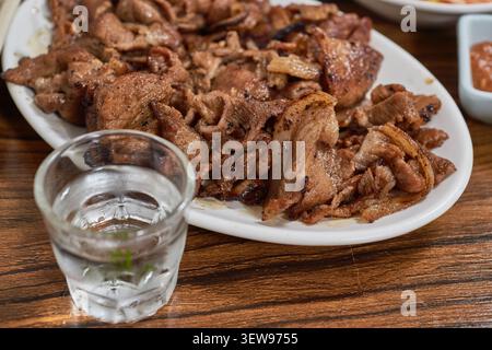 Primo piano di saporito maiale alla griglia e una bevanda alcolica limpida in un piccolo bicchiere su un tavolo di legno che mette in risalto l'autentica cultura gastronomica locale Foto Stock