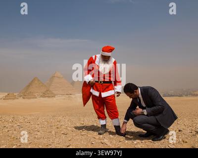 Stravagante Babbo Natale in tuta rossa assistito da un uomo d'affari inginocchiato nel deserto egiziano prima delle iconiche piramidi di Giza, ideali per la cultura incrociata Foto Stock