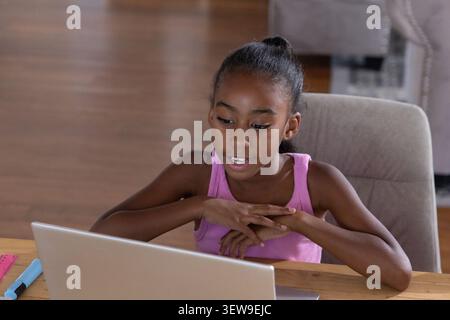 Preadolescente afro-americana seduta in canotta rosa usando il laptop a casa con righello, marcatore Foto Stock