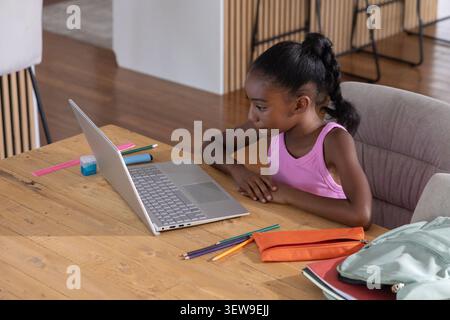 Bambina seduto a un tavolo di legno a casa utilizzando un computer portatile color argento con matite colorate e spazio per fotocopie Foto Stock