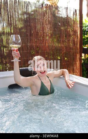 Donna spensierata che si immerge in una vasca idromassaggio all'aperto con bicchiere di vino, creando un'atmosfera di vacanza tranquilla. Foto Stock