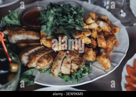 I pezzi di pollo dorato poggiano su un piatto bianco. I verdi freschi e il prezzemolo aggiungono colore e consistenza. Gli accompagnamenti di salsa suggeriscono un pasto saporito. Accogliente Foto Stock