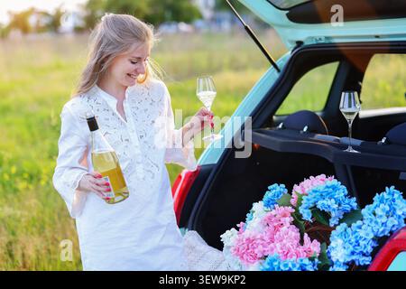 Giovane donna elegante che gusta spumante accanto a un'auto pastello piena di fiori in un prato estivo, catturando viaggi di lusso e relax in campagna Foto Stock