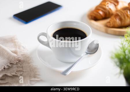 Tazza da caffè nera su tavolo bianco con croissant e smartphone in un ambiente accogliente Foto Stock