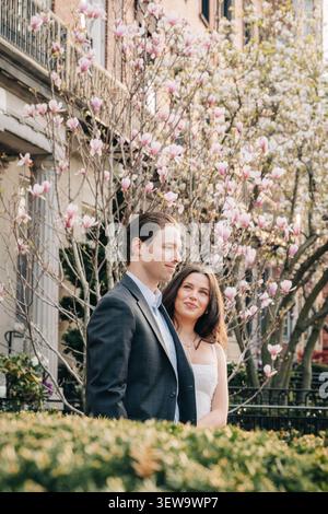 Coppia felice in posa all'aperto in una città in primavera, circondata da fiori rosa di magnolia Foto Stock
