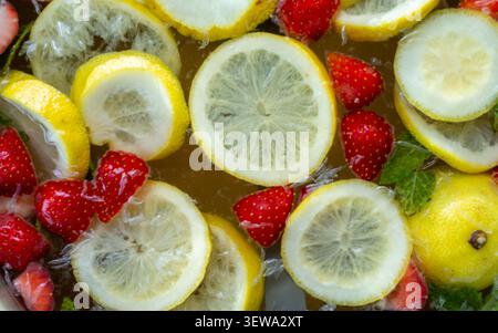 Fette di limone fluttuanti e fragole con foglie di menta in liquido raffreddato creano una rinfrescante estate luminosa, un contrasto di colori vivace, una texture lucida e uno stile alimentare fresco visto dall'alto Foto Stock