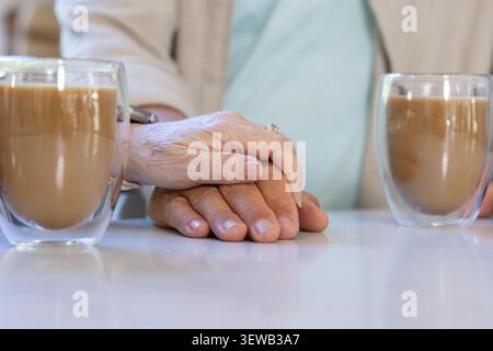 Coppie diverse che mettono le mani su un tavolo bianco lucido a casa con orologio da sposa e tazze di vetro Foto Stock