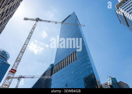Vista ad angolo basso di un impressionante grattacielo in vetro in costruzione, incorniciato da una massiccia gru a torre contro un cielo blu brillante. Rappresenta un arcipite moderno Foto Stock