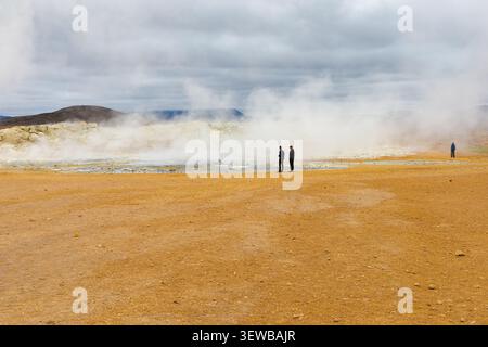 Paesaggio geotermico di Hverir in Islanda con persone che guardano un'enorme nuvola di vapore, terra minerale arancione e un cielo spettacolare nuvoloso. Foto Stock