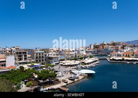 Panorama panoramico sopraelevato di Agios Nikolaos a Creta, che mostra una vivace piazza cittadina lungo il lago Voulismeni, ristoranti sul mare sotto le tende da sole, parke Foto Stock