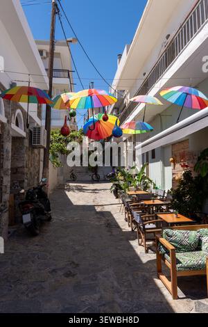 Ripresa verticale di una strada stretta e panoramica a Creta con un vivace baldacchino di ombrelli colorati sui tavoli da caffè all'aperto sotto un cielo luminoso e soleggiato. Foto Stock