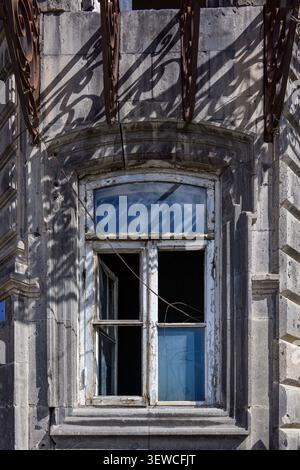 Edificio in pietra intemprata con finestra rotta. Travi arrugginite proiettano ombre su pareti vecchie. Il telaio della finestra mostra segni di decadimento Foto Stock