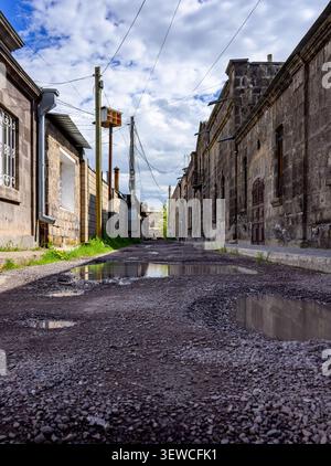 Vicolo abbandonato tra edifici in pietra. Le pozzanghere riflettono il cielo nuvoloso. Le macchie di erba ricoperte rompono la strada ghiaiosa. I fili si allungano in c.a. Foto Stock