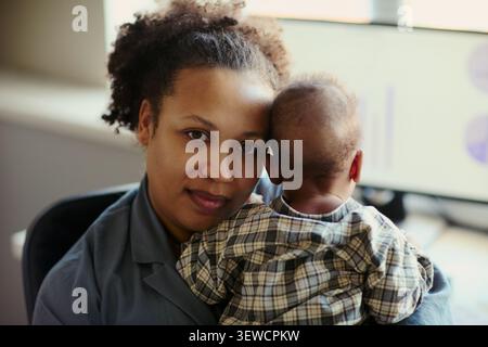 Ritratto di una giovane madre afroamericana che tiene il bambino in carica, bilanciando la cura con le responsabilità lavorative. Ideale per genitori che lavorano, politica di maternità, marketing ibrido Foto Stock