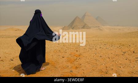 Donna araba velata in abaya nera si erge ad ammirare le piramidi di Giza attraverso il deserto egiziano nebbioso, ideale per viaggi turistici in Egitto e Medio Oriente Foto Stock
