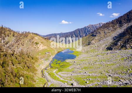 Vista aerea del lago Kareri Trek circondato da pendii rocciosi, prati lussureggianti e alberi sparsi sotto un cielo azzurro e limpido a Kareri, Himachal Pradesh, i Foto Stock