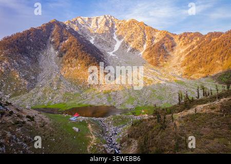 Vista aerea del lago Kareri, annidato nella catena montuosa di Dhauladhar, circondato da pendii rocciosi, prati verdi e vette innevate a Kareri, Himachal Prade Foto Stock