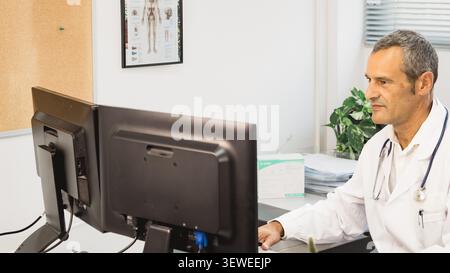 Vista laterale di un medico in cappotto bianco utilizzando un computer presso un banco della clinica, con stetoscopio, impianto e lo sfondo dello studio medico. Foto Stock