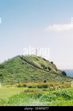 Un iconico faro bianco solitario che si erge orgoglioso sulla cima di una lussureggiante collina erbosa, sotto un cielo soffice alla luce del giorno, catturato su una pellicola vintage da 35 mm. Foto Stock