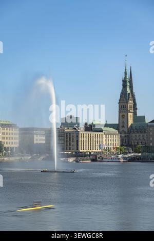 Vista del lago Alster interno illuminato dal sole di Amburgo con la torreggiante fontana Alster e una barca a remi di passaggio sul Jungfernstieg e la caratteristica torre Foto Stock