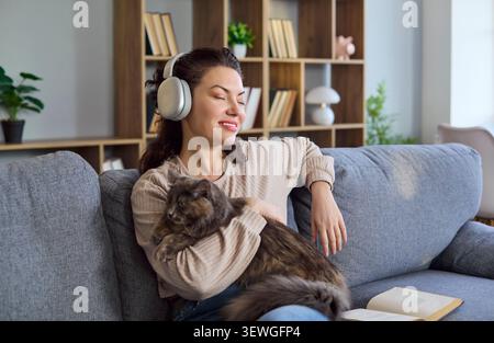 Donna che si rilassa a casa con il gatto, indossa le cuffie e ascolta la musica sul divano Foto Stock