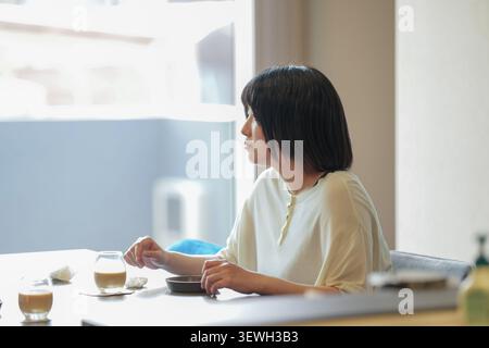 Una luce soffusa penetra attraverso una finestra. La donna siede tranquillamente a un tavolo. Il suo sguardo si sposta verso il mondo esterno. Le tazze da caffè riposano nelle vicinanze, intatte. La r Foto Stock