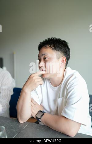 Il giovane si siede con cura, mano vicino alla bocca. Indossa una semplice T-shirt bianca e uno smartwatch. La luce naturale soffusa inonda gli interni minimalisti. Il suo gaz Foto Stock