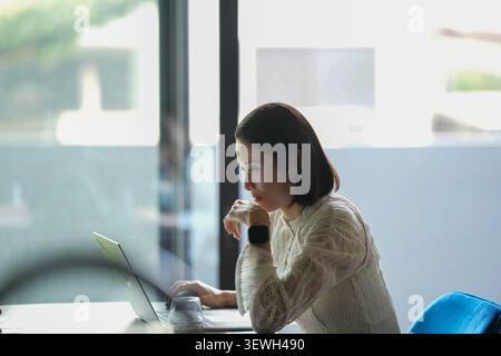 La donna siede silenziosamente, concentrata sullo schermo di un notebook. Sorseggia caffè, occhi fissi sul lavoro. La luce soffusa filtra attraverso l'ampia finestra dietro. Calma, premurosa Foto Stock