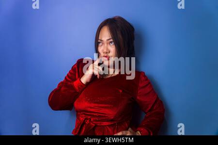 Giovane donna asiatica che tocca il mento con un'espressione premurosa su uno sfondo blu, simboleggia il pensiero, il dubbio e il processo decisionale. Foto Stock