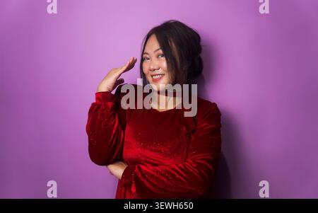 La giovane donna asiatica che indossa un abito in velluto rosso sorride mentre posa con una mano sollevata accanto al viso su uno sfondo viola dello studio. Foto Stock