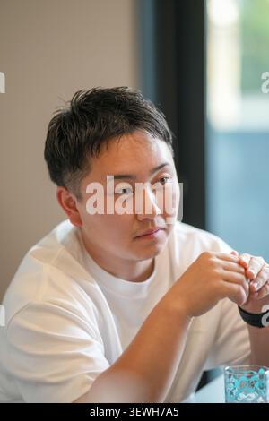 Giovane uomo in maglietta bianca seduto con cura. Il suo sguardo è lontano, mostrando una contemplazione tranquilla. Le dita poggiano vicino a un bicchiere, suggerendo una pausa o un riflesso. Foto Stock