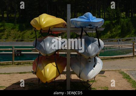 Un'ampia scelta di kayak e canoe in plastica multicolore conservate su scaffali metallici presso una stazione di noleggio vicino a un panoramico lago di montagna. Una densa pineta verde Foto Stock