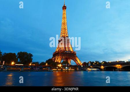 Parigi, Francia - 18 novembre 2025 : la Torre Eiffel illuminata si illumina d'oro contro un cielo blu scuro al crepuscolo a Parigi, riflesso nella calma Senna belo Foto Stock