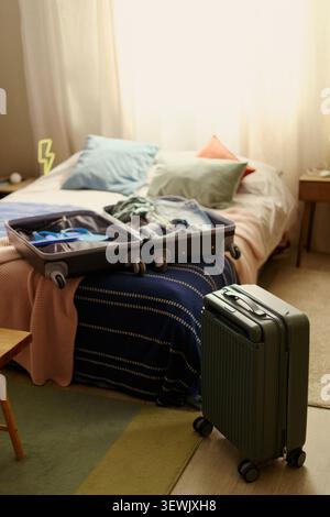 Immagine verticale di valigia aperta appoggiata sul letto, piena di vestiti e oggetti da viaggio, con bagagli rotanti nelle vicinanze, suggerendo di pianificare il viaggio. Prenotazione vacanza di coppia, annunci di viaggio, marketing ospitalità Foto Stock