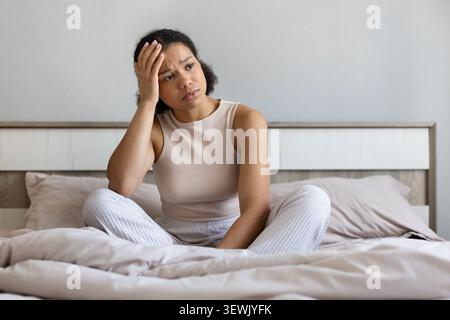Donna nera preoccupata seduta sul letto con una mano sulla fronte Foto Stock