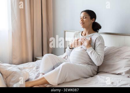 Donna nera incinta seduta sul letto che pratica esercizi di respirazione calma Foto Stock