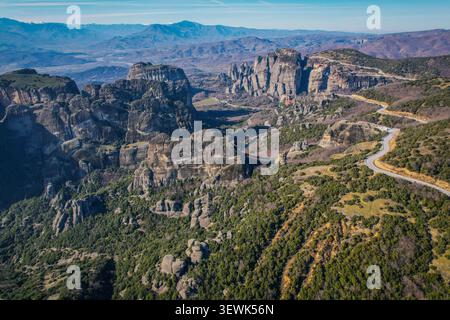 Un'ampia vista aerea cattura le suggestive e antiche formazioni rocciose di Meteora, Grecia, ricoperte di vegetazione verde. Una tortuosa strada asfaltata Foto Stock