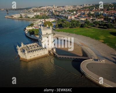 Un grandangolo aereo cattura la torre di Belém situata sul fiume Tago con una spiaggia di sabbia e un passaggio pedonale curvo adiacente all'erba verde. L'im Foto Stock