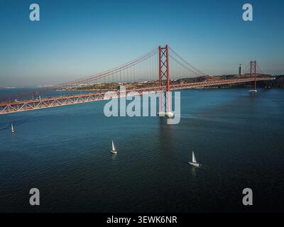 Il ponte 25 de Abril attraversa il fiume Tago a Lisbona, in Portogallo, con il santuario di Cristo Rei visibile sulla riva opposta. Tre barche a vela bianche Foto Stock