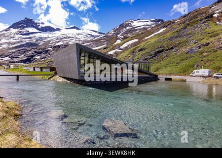 Trollstigen, Norvegia 5 - 2026 giugno: Moderno edificio galleggiante sopra il limpido ruscello di montagna. Le cime innevate si innalzano dietro verdi e aspre colline. Il cielo è più luminoso Foto Stock